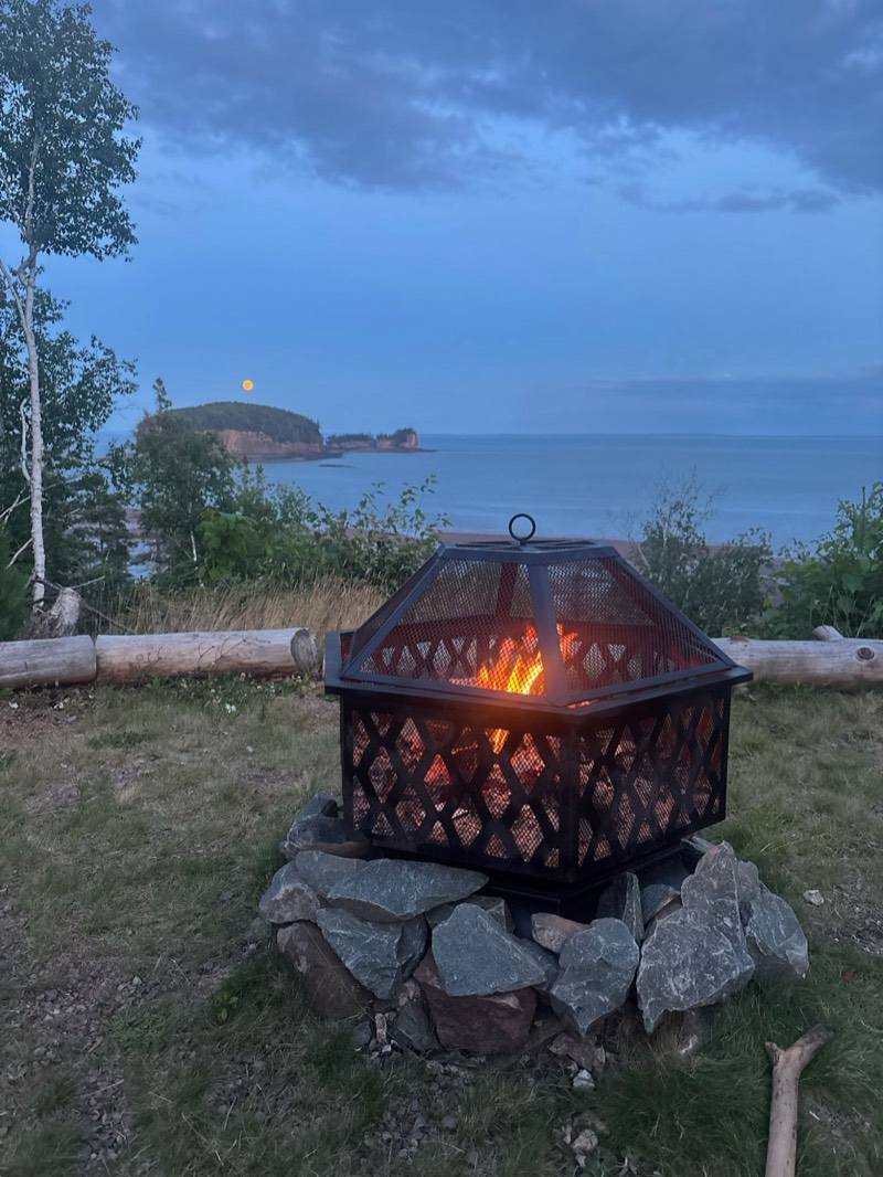 A crackling fire pit at dusk with the moon rising over the Two Islands on the Bay of Fundy