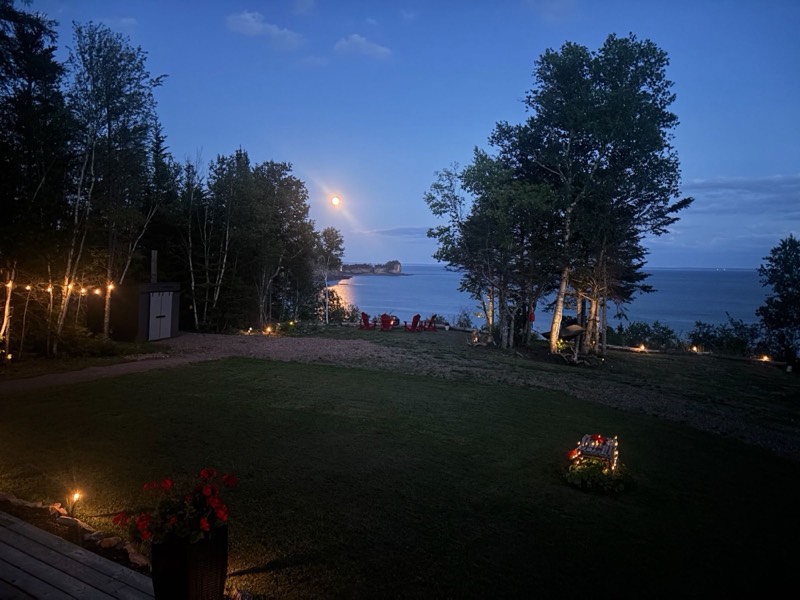 The cottage lawn at night with string lights, red chairs and the moon reflecting on the Bay of Fundy